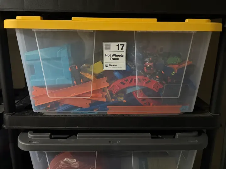 A clear storage bin with a yellow lid on a black shelf, a Muninn label facing out: a QR code, a big 17, “Hot Wheels Track” and the Muninn raven. Through the plastic, orange and blue toy car track and a heap of toy cars. Another bin sits on the shelf below.